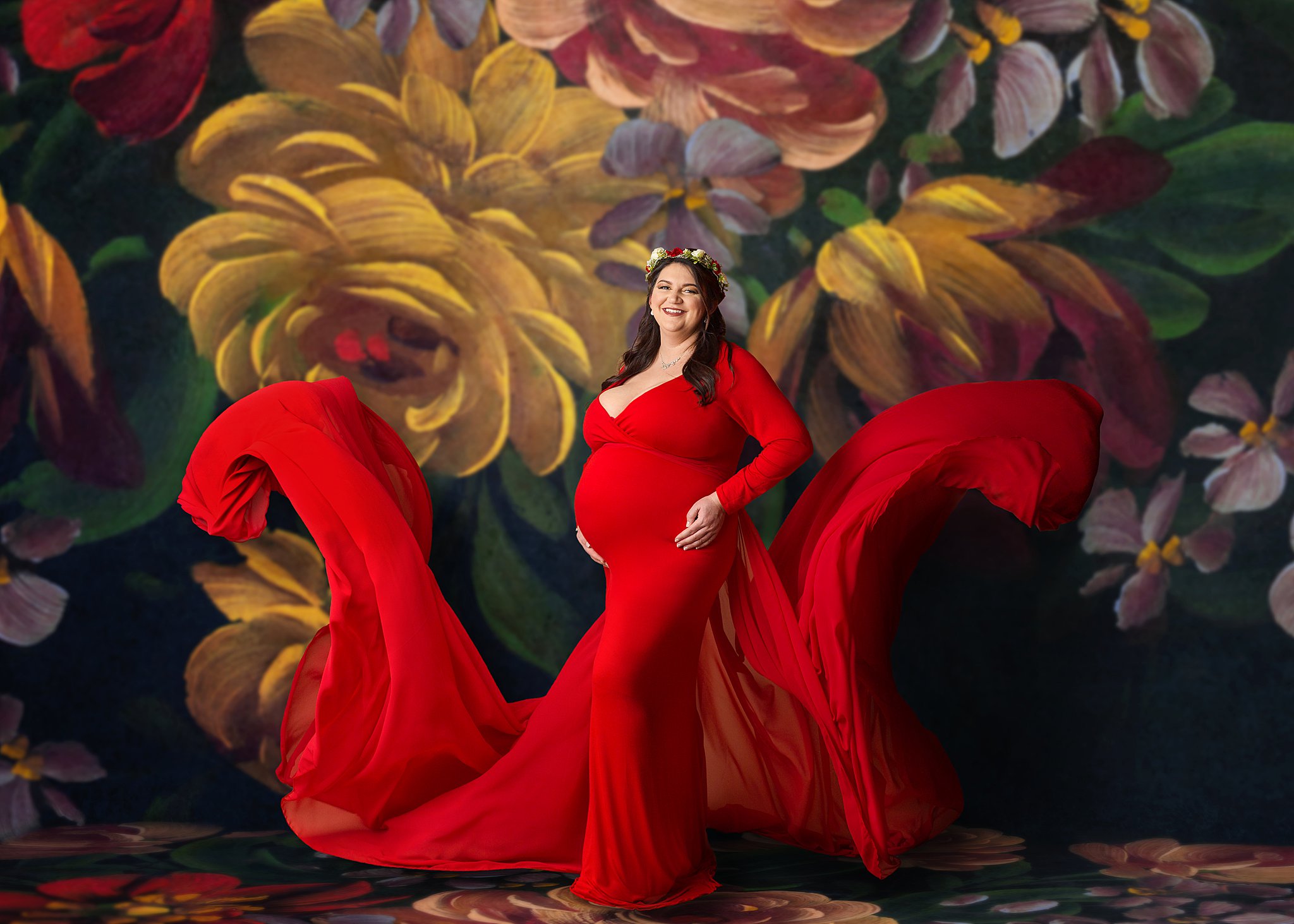 Pregnant woman posing on a floral backdrop in a red flowy gown from Moline Illinois.